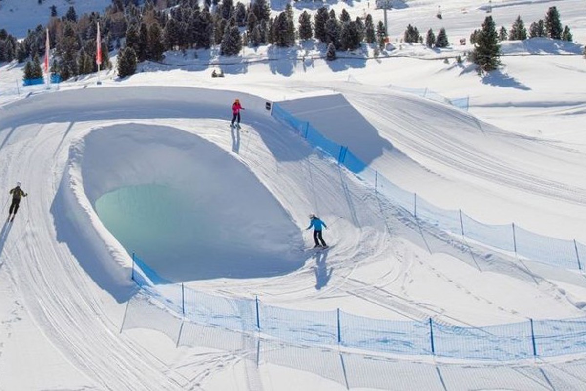 Tre skidåkare åker i en snöpark med en stor doserad kurva runt en snötunnel, omgiven av skyddsnät i ett alpint vinterlandskap.
